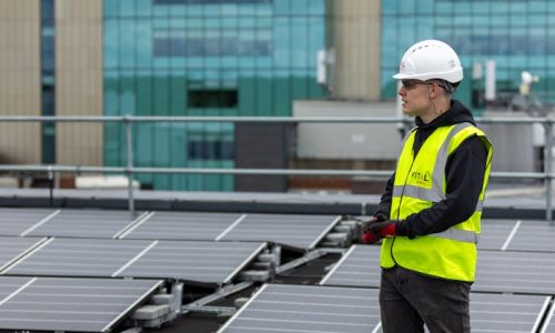 A worker in a safety vest and helmet inspects solar panels on an urban rooftop, promoting renewable energy.